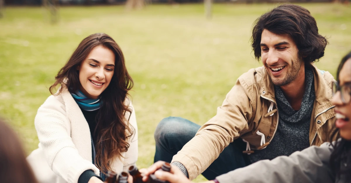 Positive Wellbeing Psychology Melbourne | Social Anxiety vs. Shyness: How to Tell the Difference Group of young friends smiling and clinking glass bottles together while sitting on the grass outdoors