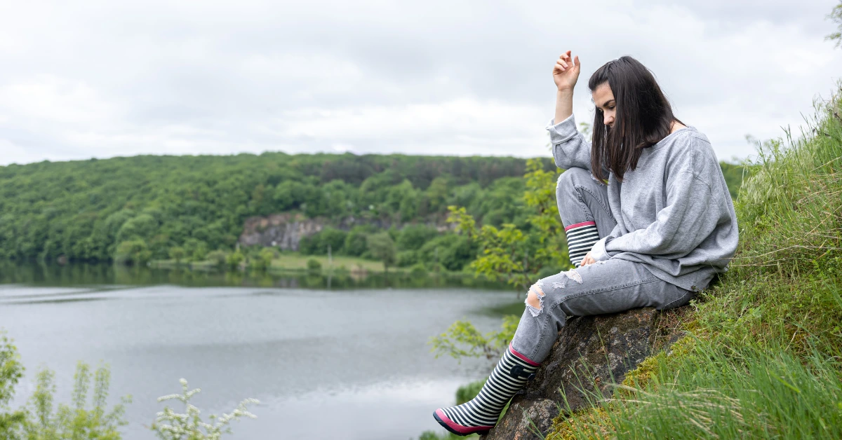 Woman in a gray sweatshirt and striped rain boots sitting on a grassy hill overlooking a lake