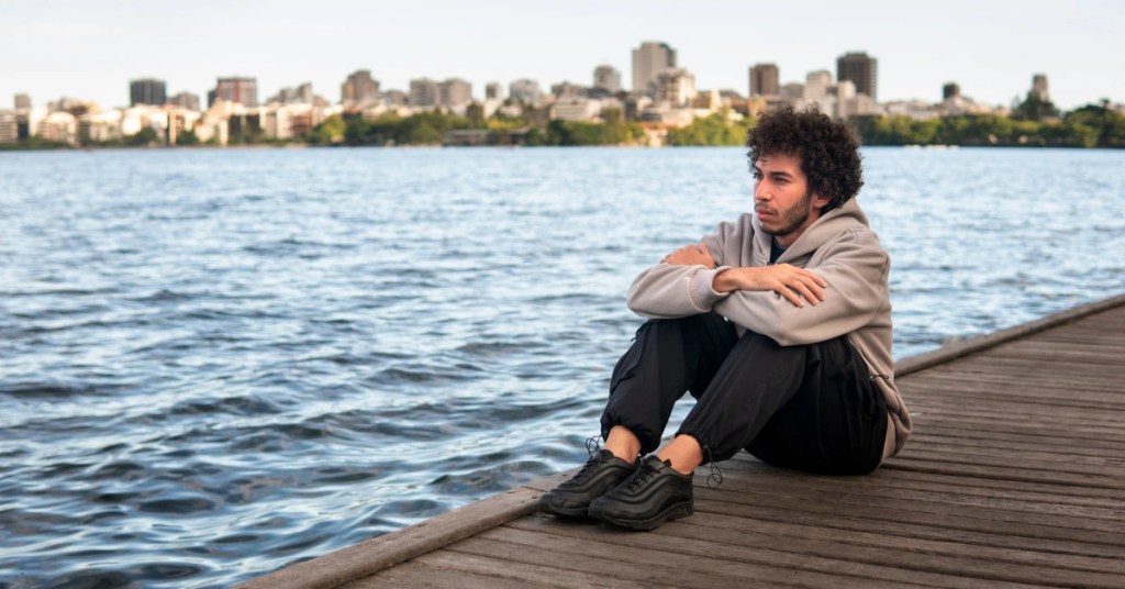 Young man with curly hair sitting on a wooden dock with arms crossed looking out over water toward a city skyline