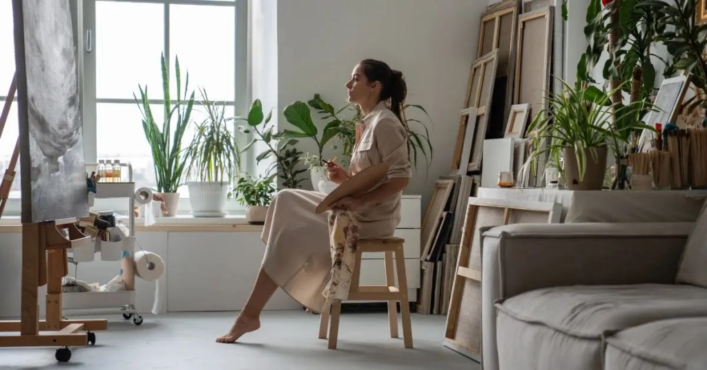 Positive Wellbeing Psychology Melbourne | Low Confidence in High Achievers: The Hidden Block Behind Success Female artist sitting on a wooden stool holding a palette in a bright art studio filled with plants